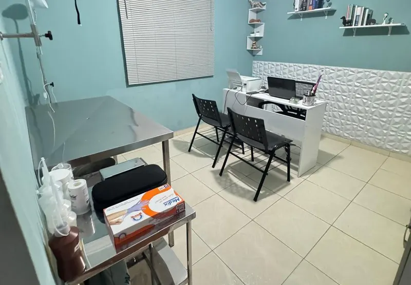 Consultório com mesa de exame de inox, mesa de atendimento e prateleiras com livros
