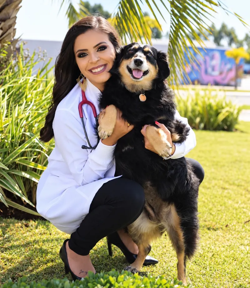 Dra. Ingrid Zaquine de jaleco e estetoscópio, sorrindo e abraçando um cachorro preto e caramelo no jardim